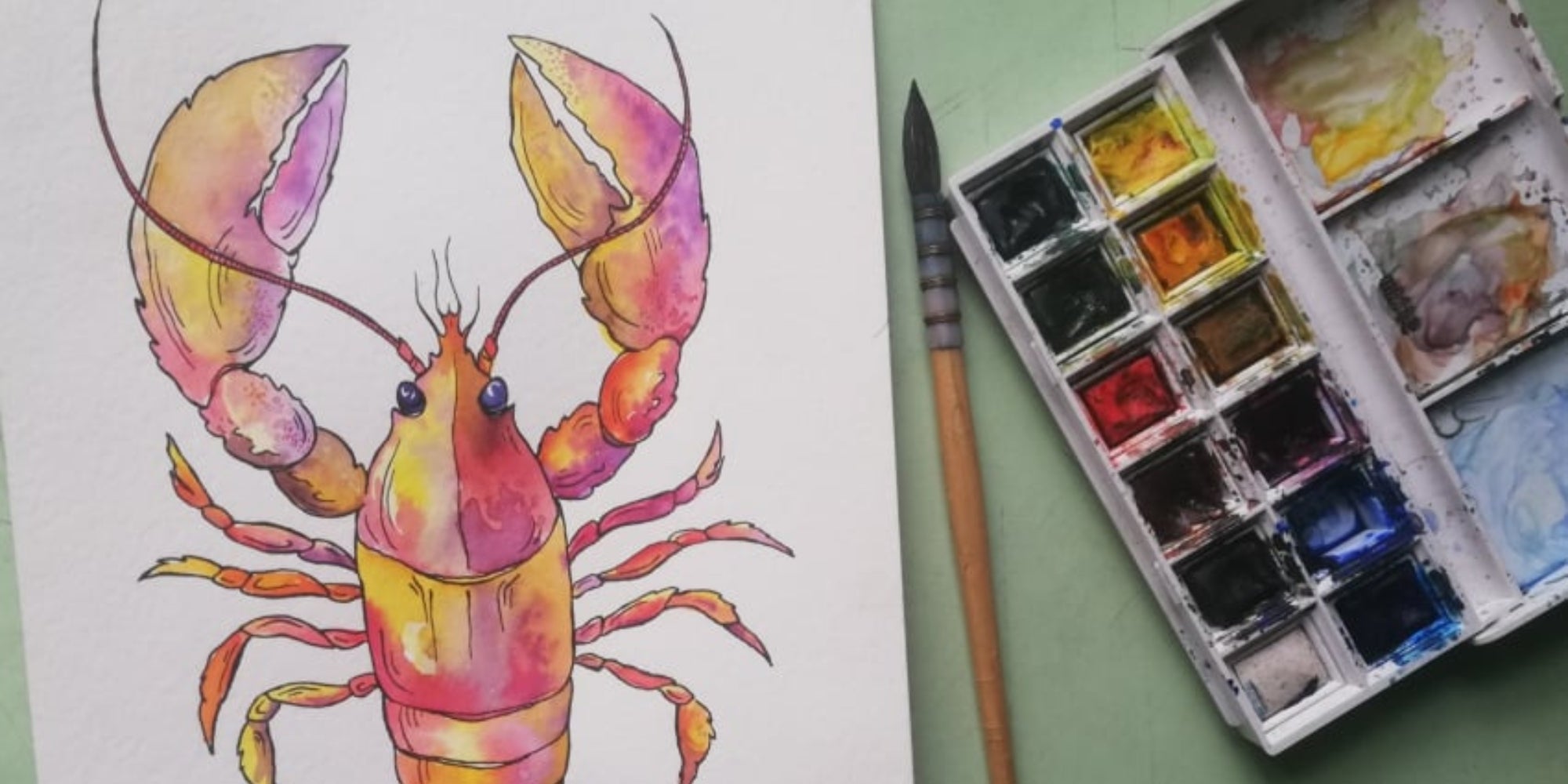 A colorful watercolor painting of a lobster in pink, yellow, and purple hues, sitting next to a used watercolor palette and brush.
