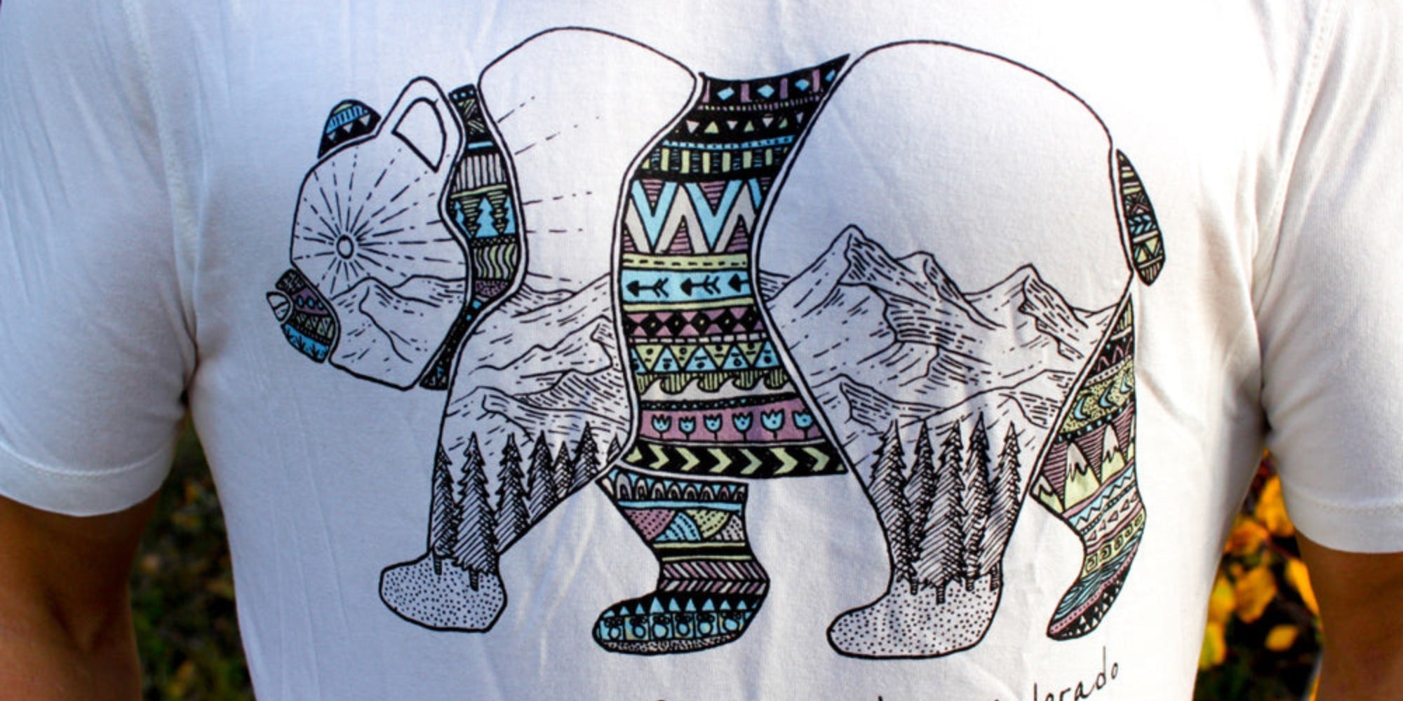 A white t-shirt featuring a stylized silhouette of a bear, filled with intricate geometric patterns and a mountain landscape illustration.