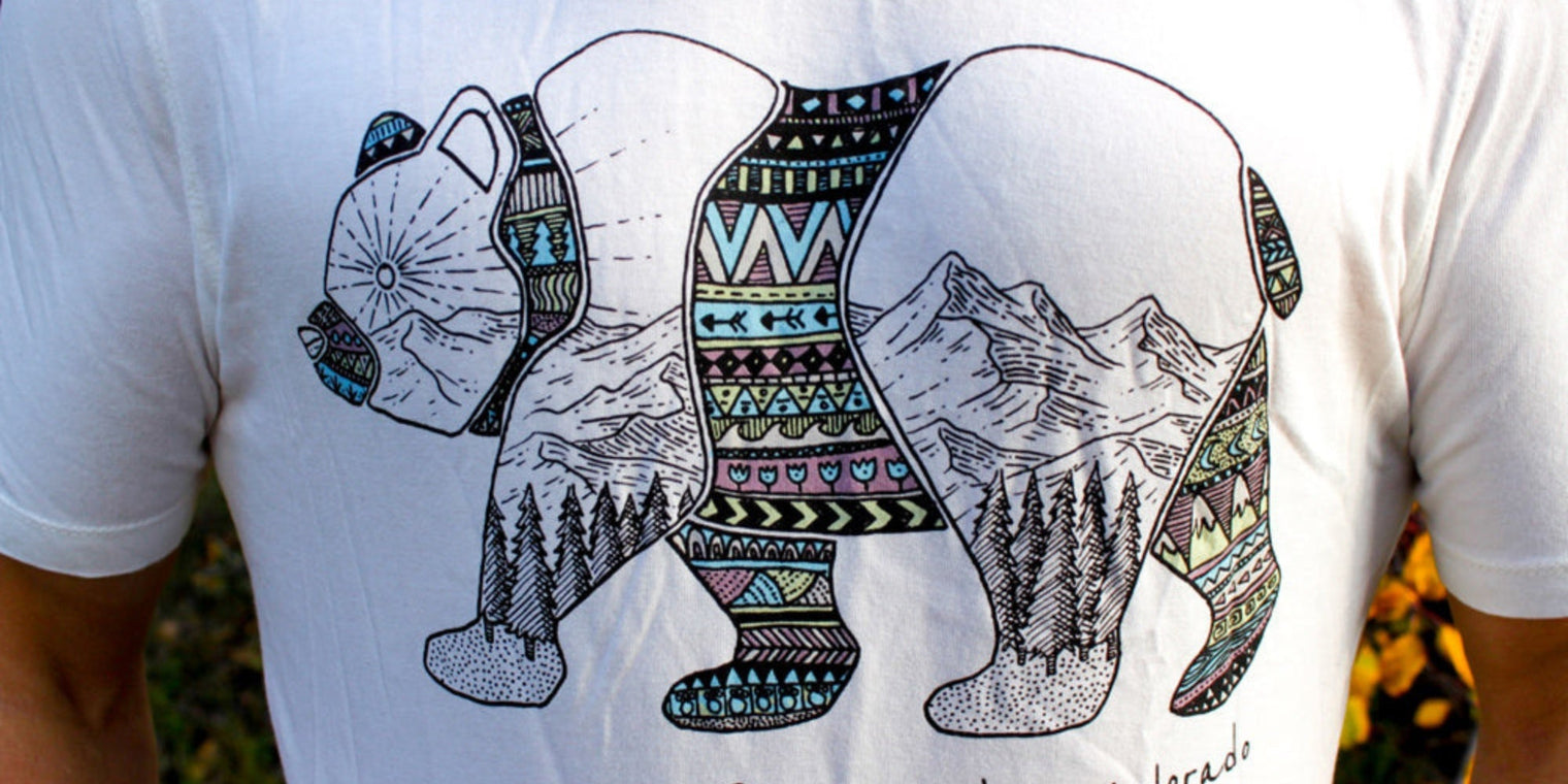 A white t-shirt featuring a stylized silhouette of a bear, filled with intricate geometric patterns and a mountain landscape illustration.